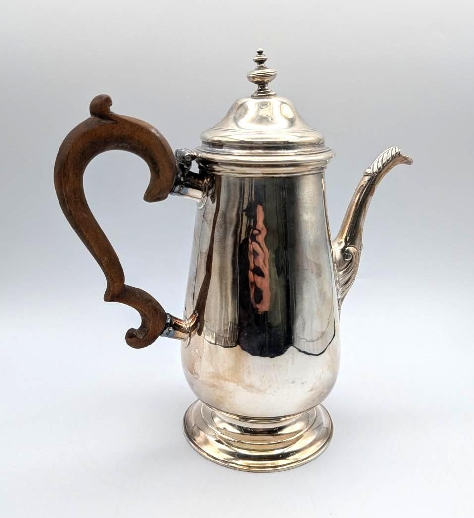A silver coffee pot with a curved wooden handle, a lid with a finial, and a long spout, set against a plain background.