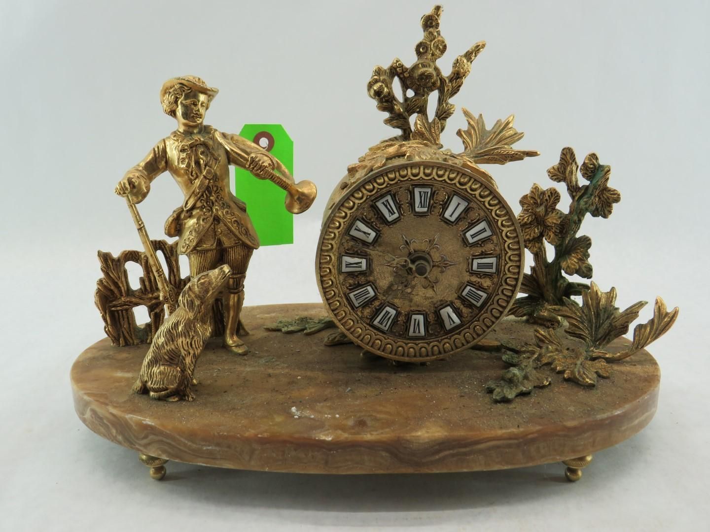 Antique gold clock with hunter, dog, and foliage on a circular base.