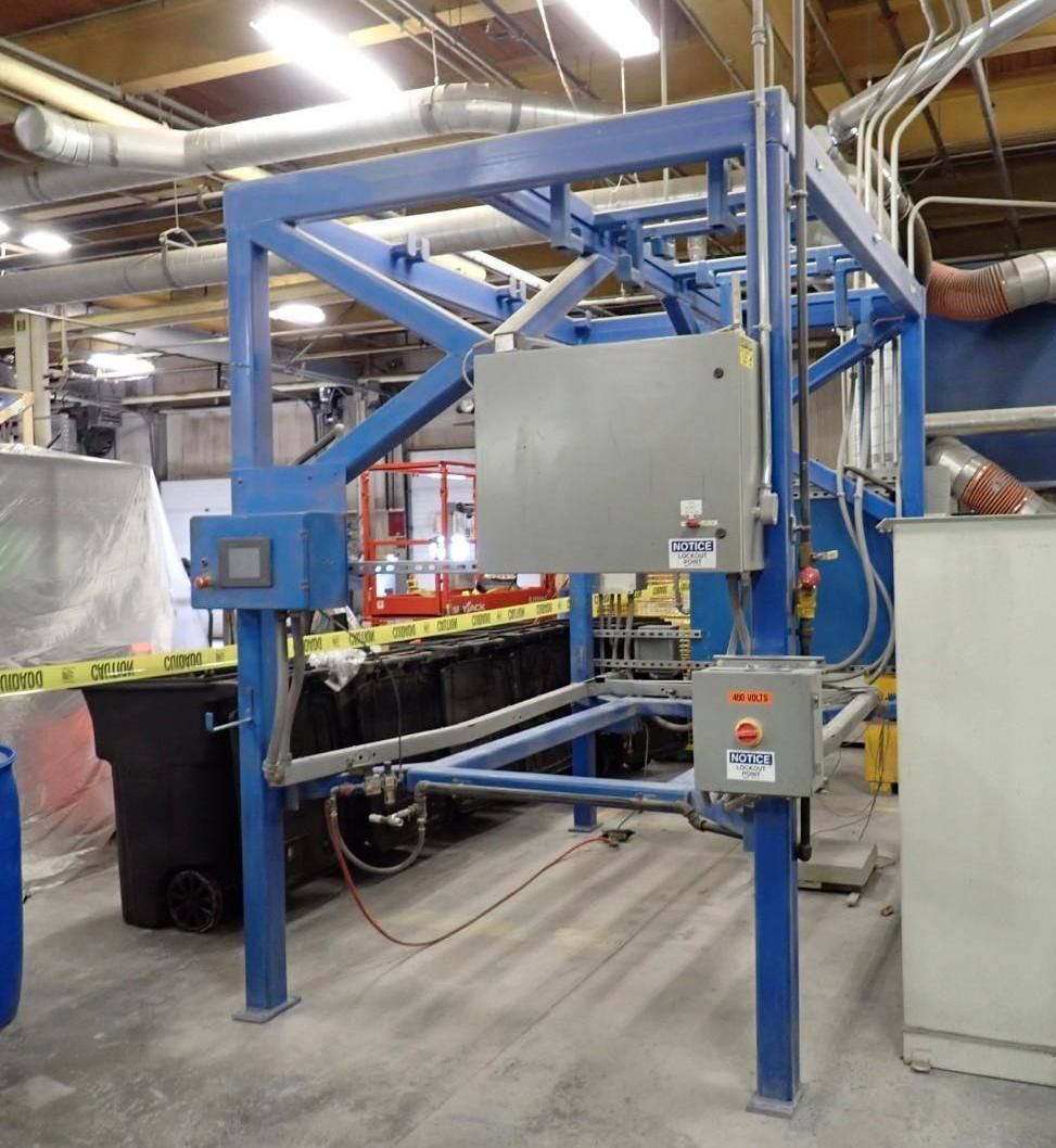 Blue metal industrial gantry over dark bins, with electrical boxes and control panel in a factory setting.