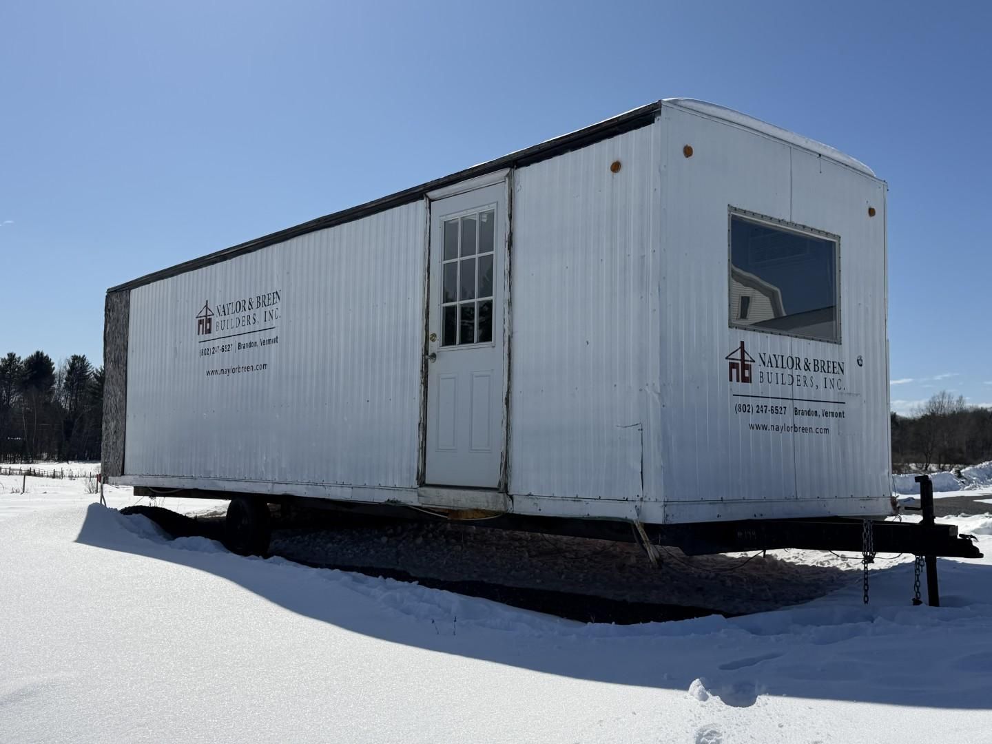White construction trailer on a snowy ground against a blue sky. A door and window are visible.