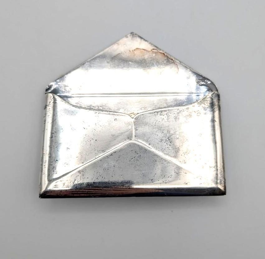 A small, silver-colored, metallic object shaped like a classic folded envelope, centered on a plain light grey background.