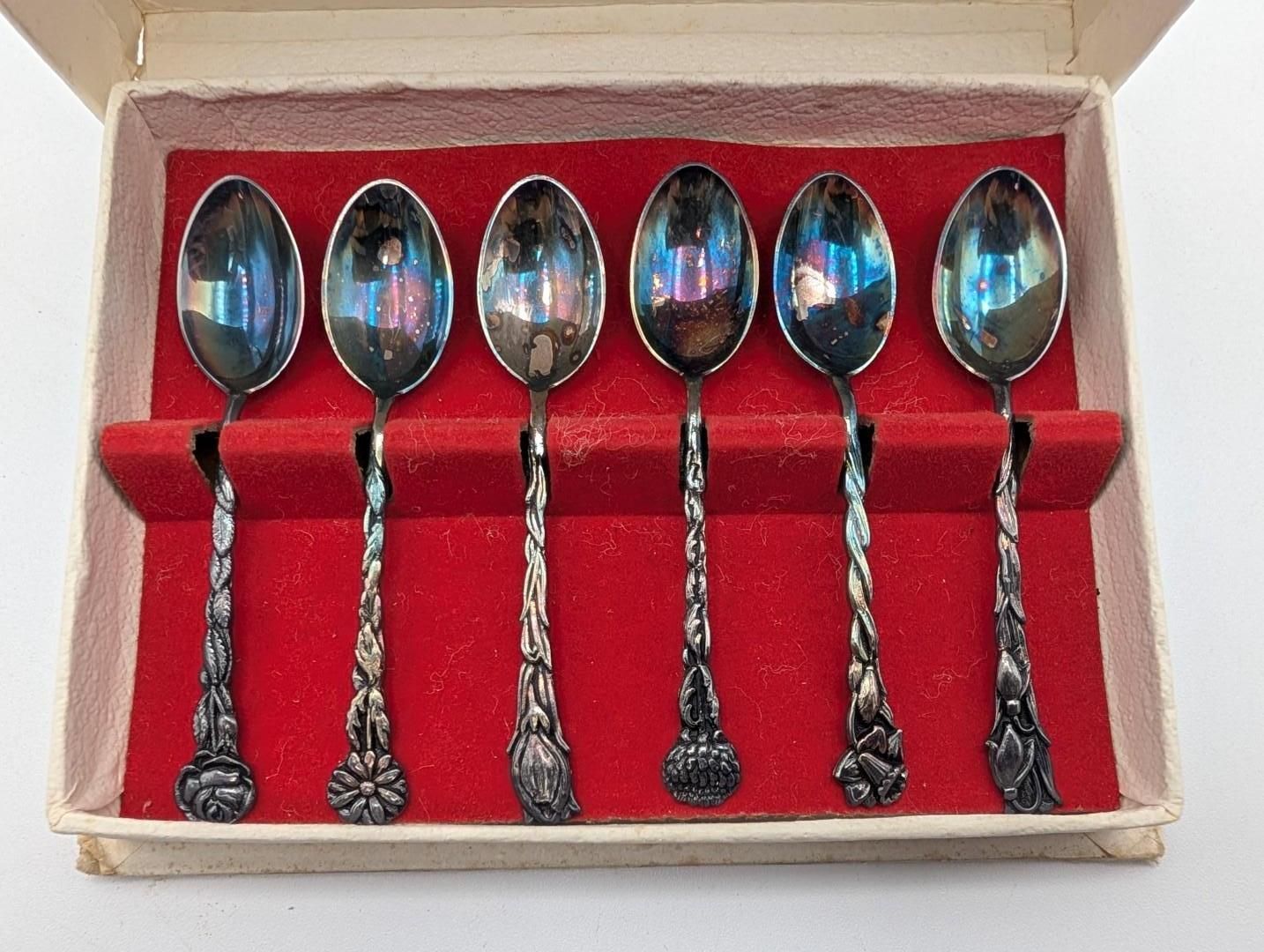 A set of six silver-toned spoons with ornate, twisted handles, displayed in a red-lined, off-white storage box.