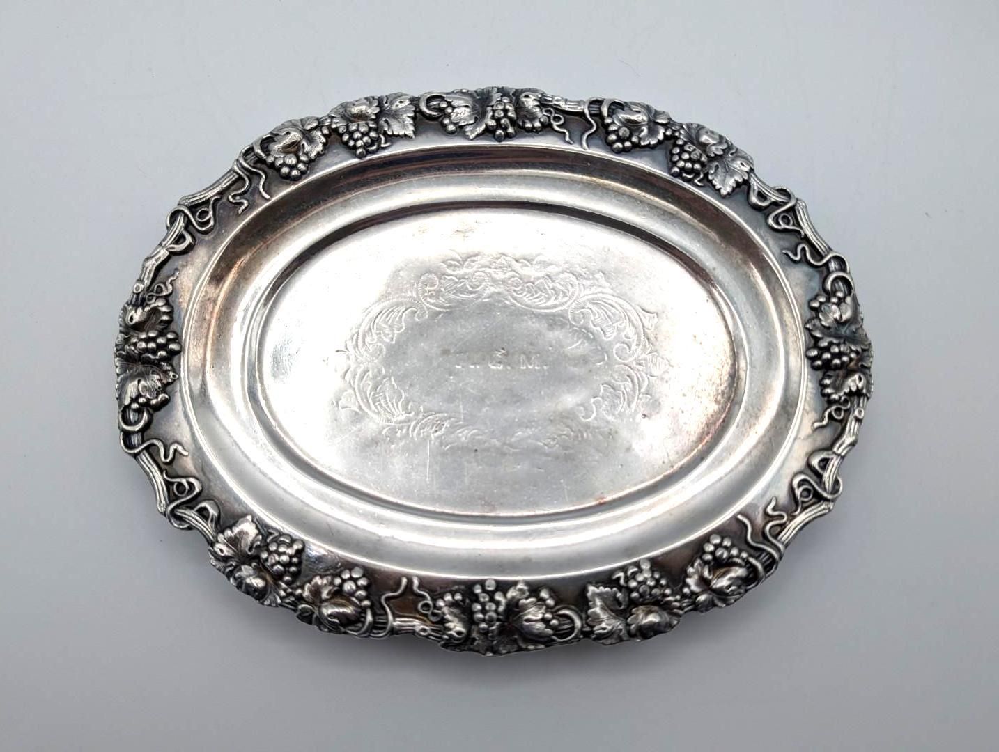 An ornate, oval silver tray with an intricate grape and vine motif along the rim and a decorative engraving in the center.