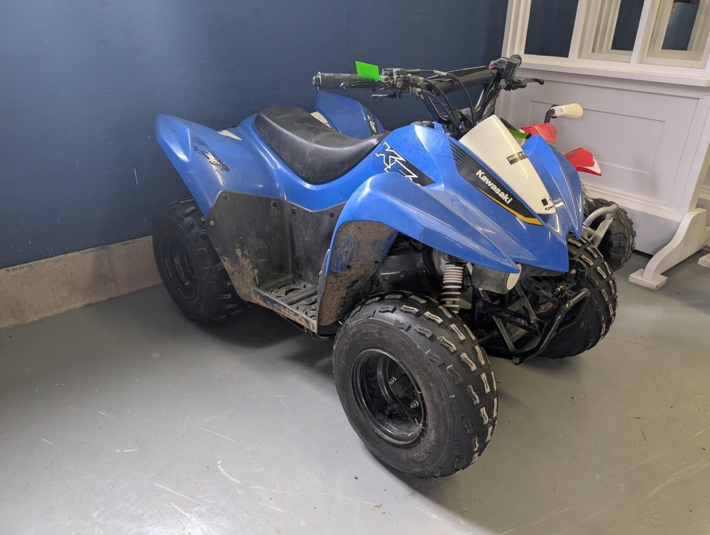 Blue ATV parked indoors near a white window, with black tires and seat, on a gray floor.