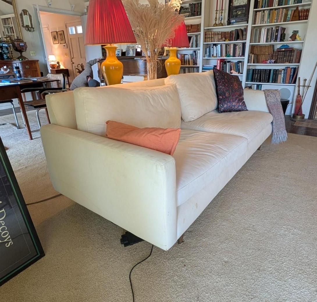 A cream-colored sofa sits in a living room with bookshelves, two yellow lamps, and a patterned throw pillow.