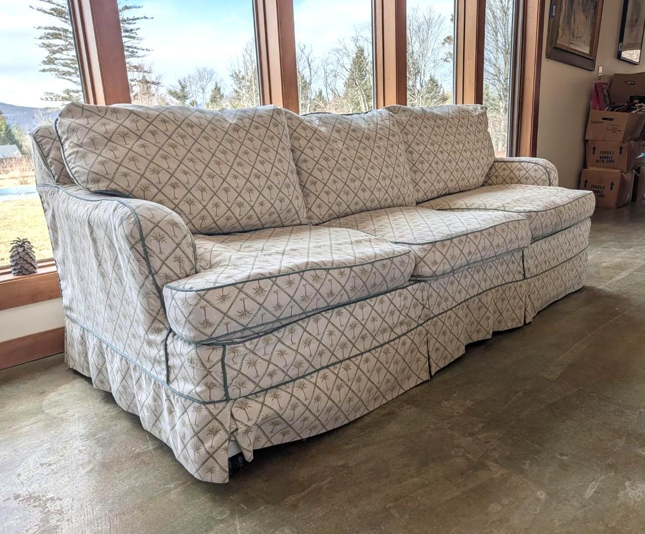 A three-cushion sofa with a blue and white diamond-patterned fabric and a pleated skirt, positioned in a sunny room.