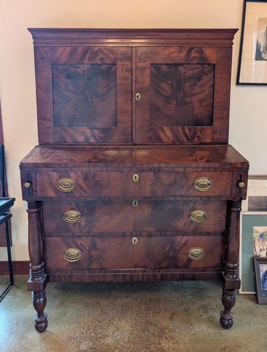 A tall, dark wood mahogany secretary desk with a hutch, brass hardware, and turned legs, standing in a room.