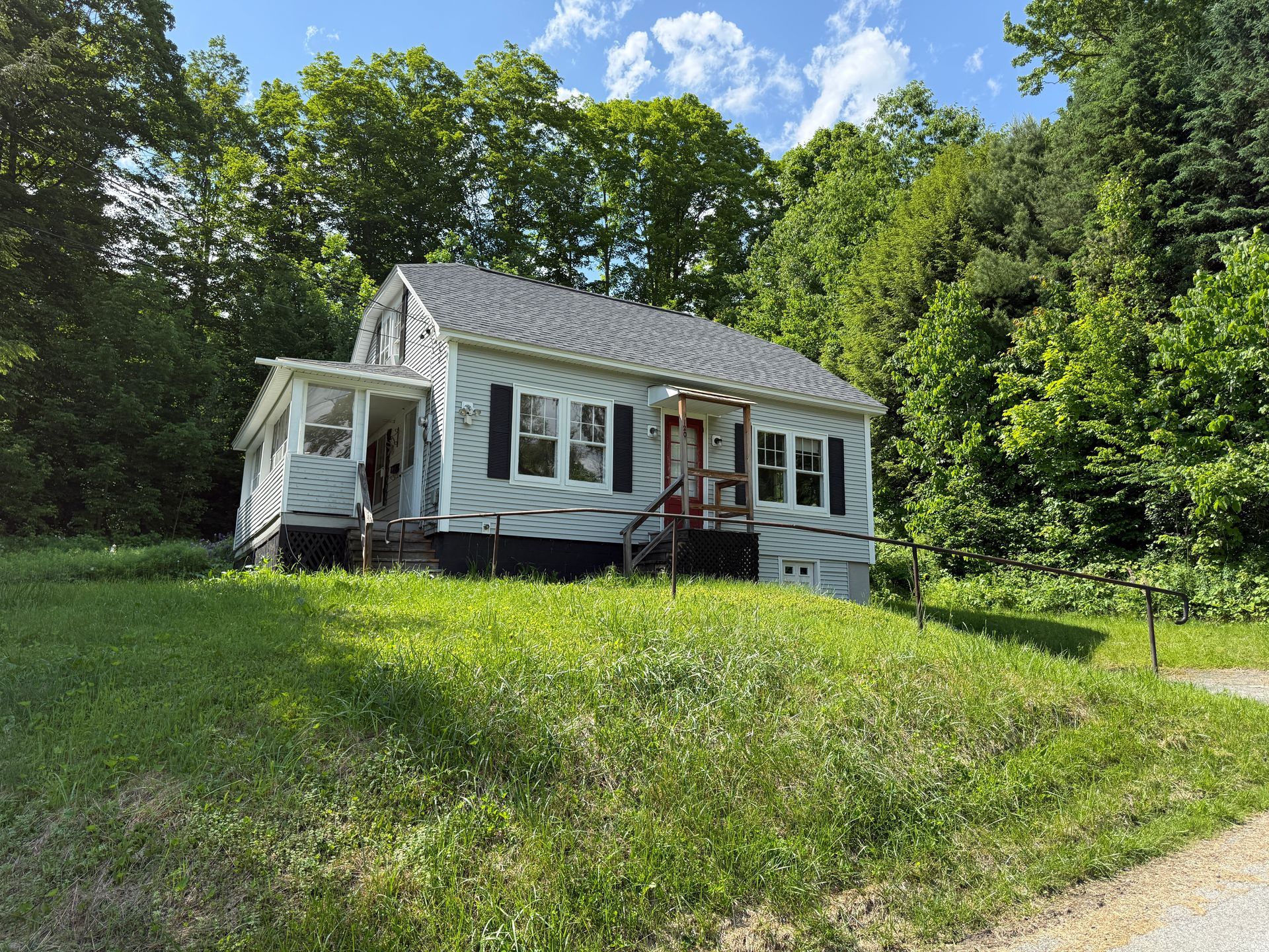 White cottage with porch on a grassy hill surrounded by trees and greenery under a bright sky