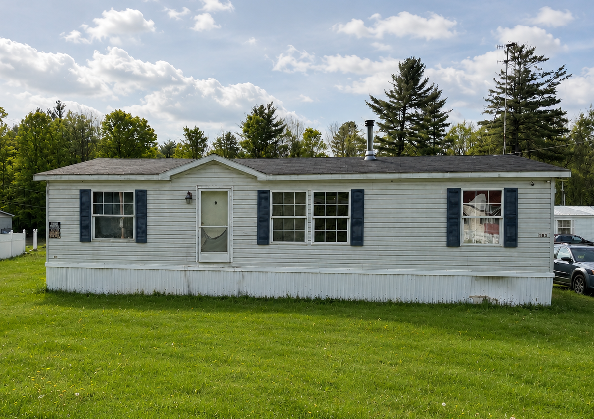 White single-wide mobile home with gray shutters on a grassy lot under a cloudy sky