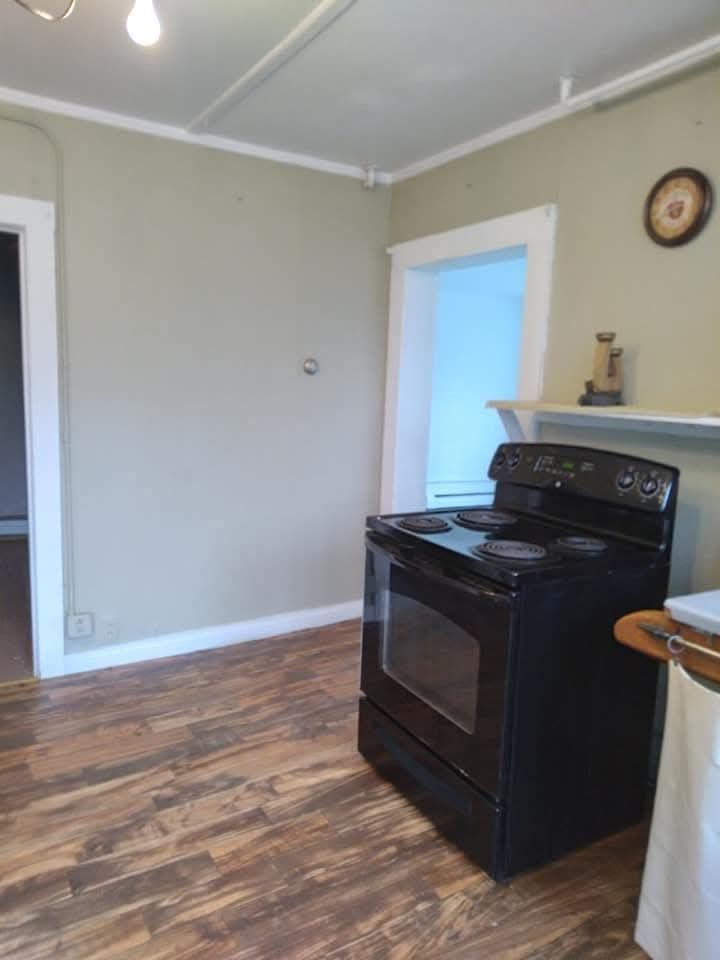 A black electric stove sits against a light-toned wall in a room with wood-patterned flooring and a doorway to the left.