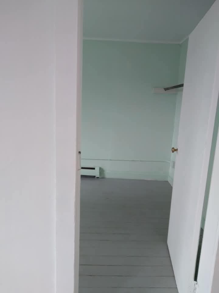 An open white door reveals a sparsely furnished room with light green walls and painted gray wooden floorboards.