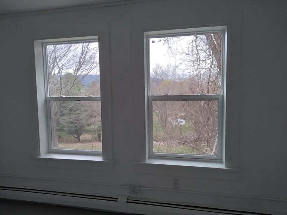 Two white-framed windows set into a plain white wall, looking out onto a view of trees and a distant hillside.