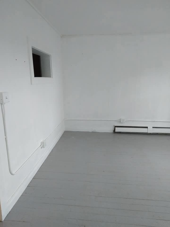 A corner of an empty room with white walls, gray wood floors, a small rectangular window, and a wall-mounted heater.