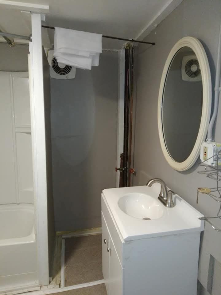 A small bathroom with a white vanity sink, an oval mirror, and a shower stall to the left with towels hanging above.