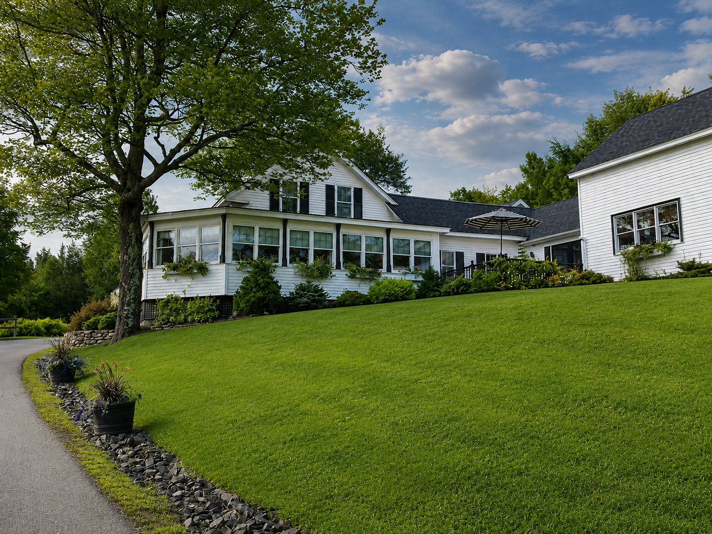White house with a front porch on a green hillside, bordered by trees and a curved stone edge.