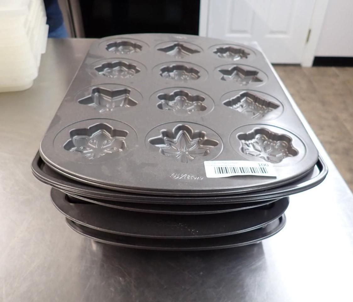 A stack of dark metal baking pans on a stainless steel counter, each designed with twelve distinct leaf-shaped molds.