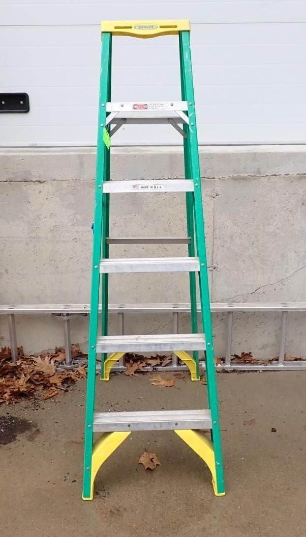 A green and yellow fiberglass stepladder with five steps standing on a concrete surface against a wall.