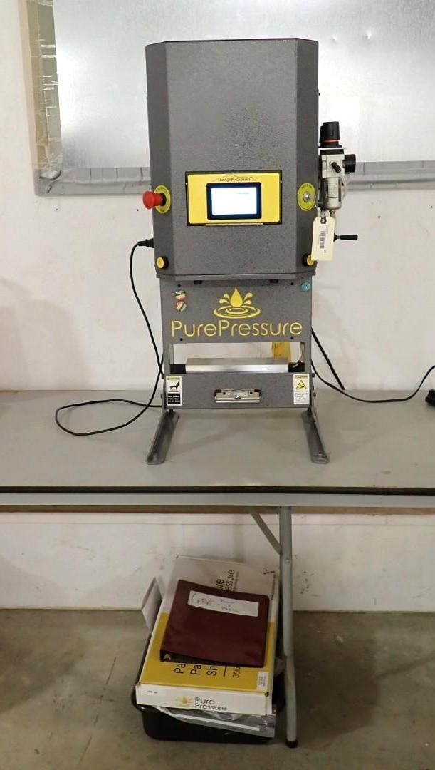 A grey PurePressure rosin press machine sits on a white folding table with a manual and supplies stored underneath.