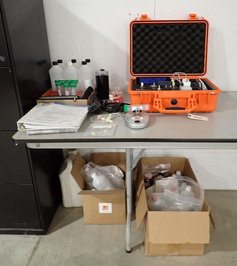 Scientific field equipment including an open orange case, bottles, and testing supplies on a folding table and in boxes.