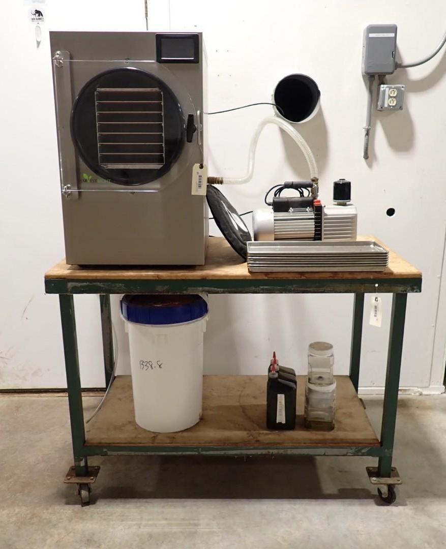 A freeze dryer and vacuum pump sit on a metal utility cart against a white wall with electrical equipment.