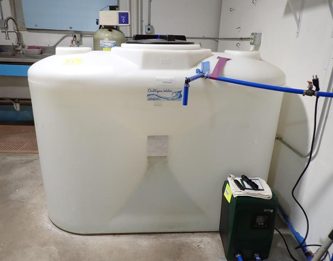 A large, white, rectangular plastic water storage tank sits in a room next to a pump and a blue pipe system.