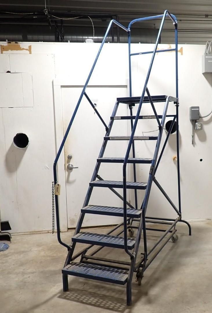 A blue industrial rolling ladder with seven steps and side handrails, standing on a light concrete floor.