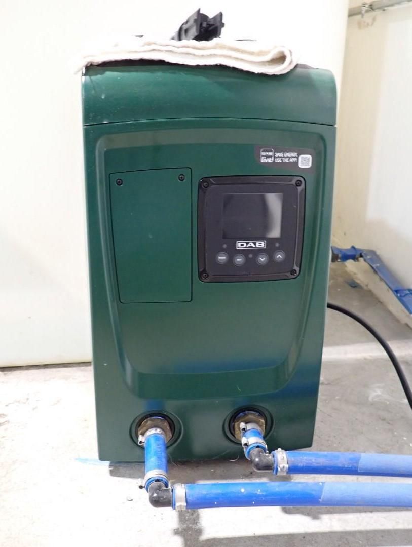 A green, rectangular variable speed pool pump with a digital display panel, connected to blue pipes on a concrete floor.