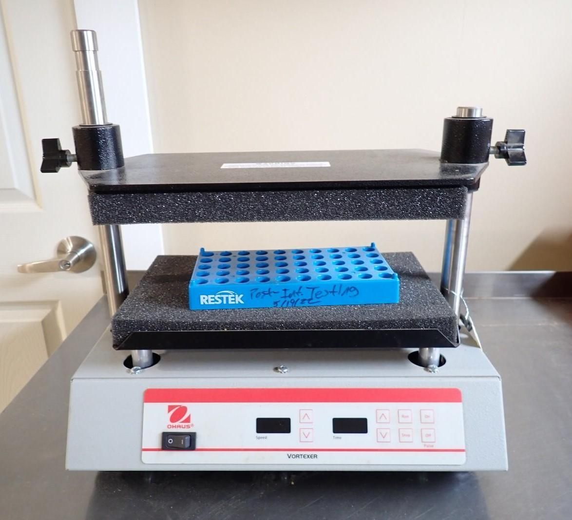A laboratory digital vortex shaker with a blue test tube rack sitting on its shaking platform.