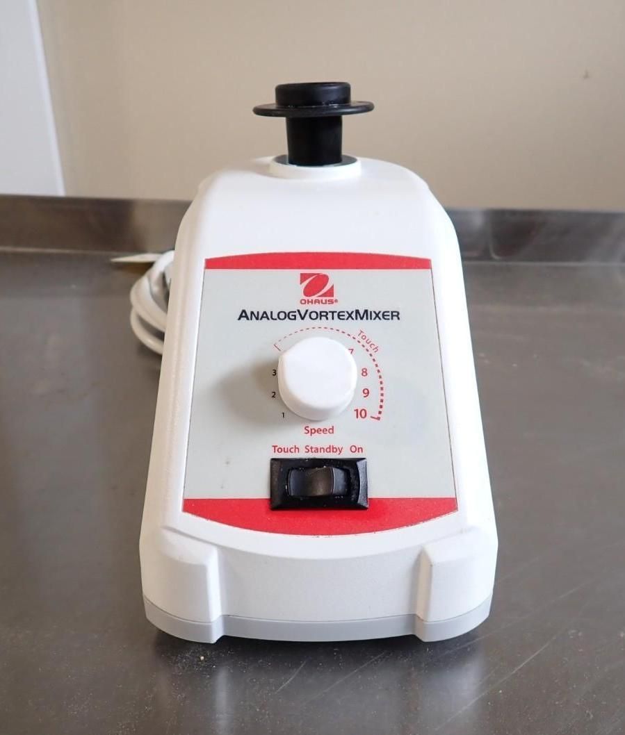 A white Ohaus lab vortex mixer sits on a metal surface, featuring a speed control dial and a power switch.