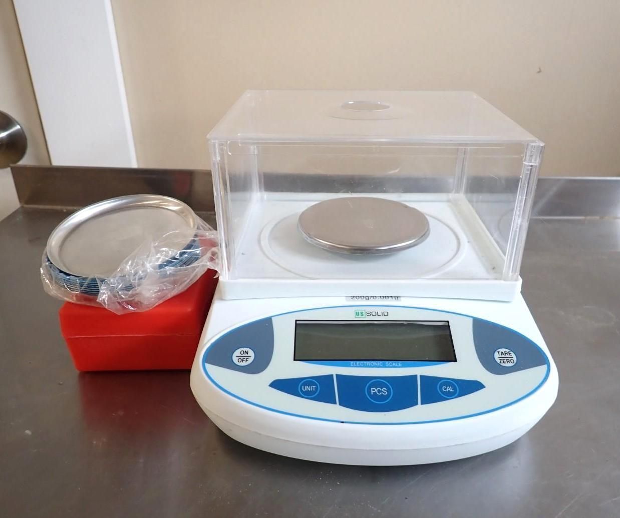 A digital precision balance with a clear protective cover sits next to a small metal weighing tray on a stainless surface.