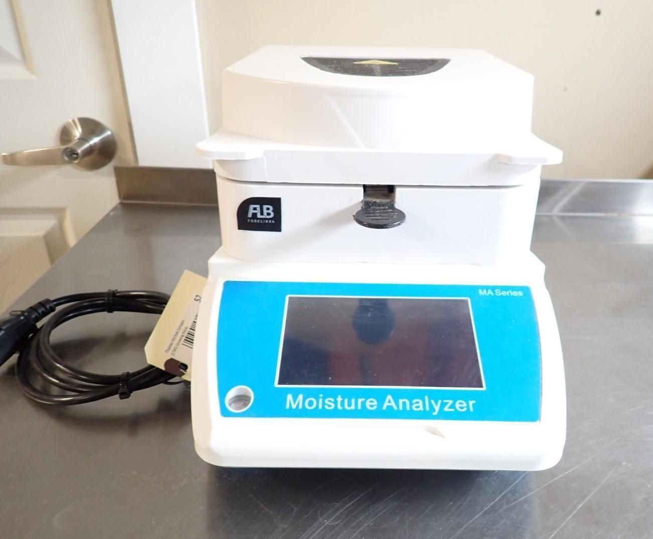 A white FLB Moisture Analyzer sits on a metal table next to its black power cord, featuring a blue digital display.