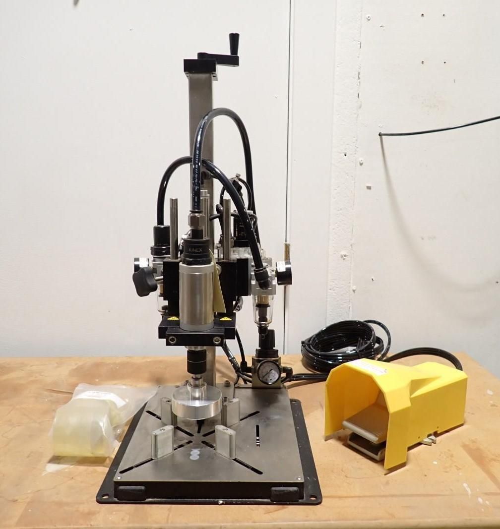 A pneumatic desktop press machine with a vertical stand, circular metal base, and a yellow foot pedal on a wooden surface.