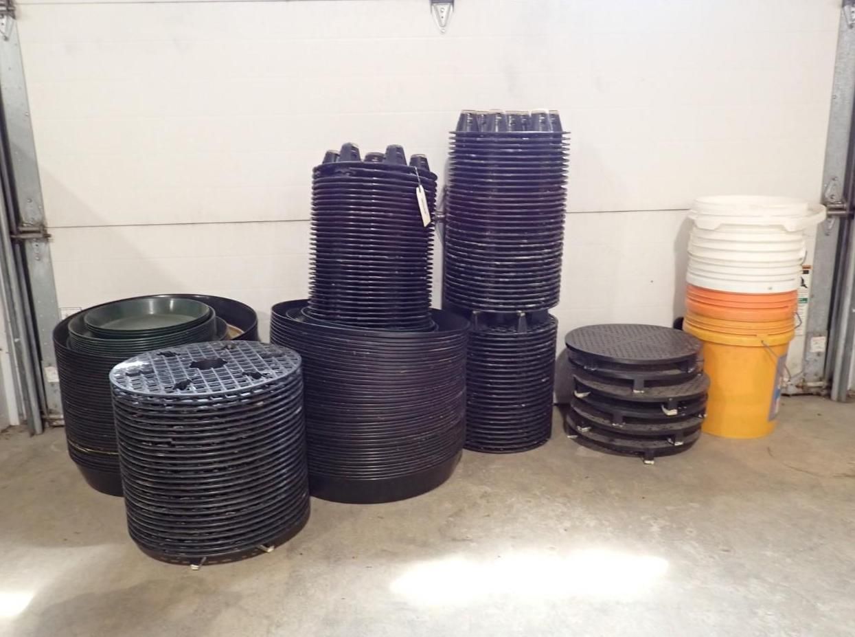 Stacks of black plastic plant nursery pots, circular trays, and a column of white and yellow buckets on a garage floor.
