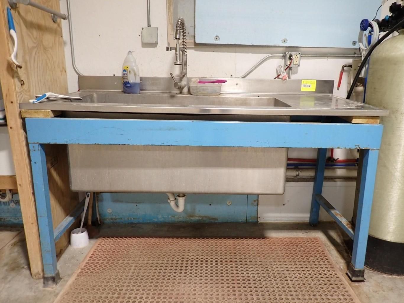 A stainless steel utility sink set in a blue metal frame in a basement, with a faucet, cleaning supplies, and floor mat.