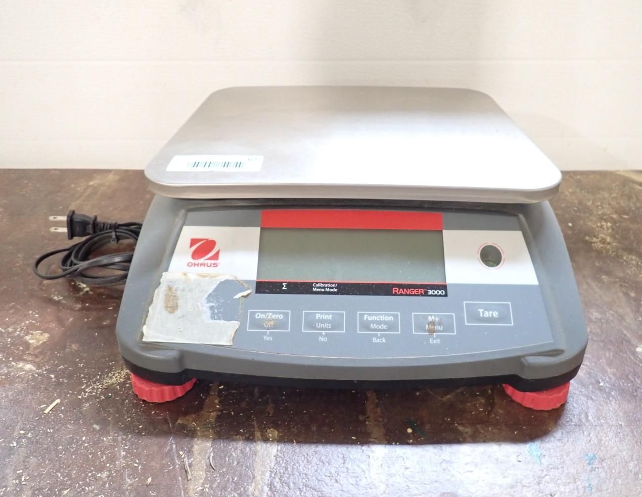 A grey Ohaus digital bench scale with a stainless steel platform, power cord, and red leveling feet on a wooden surface.