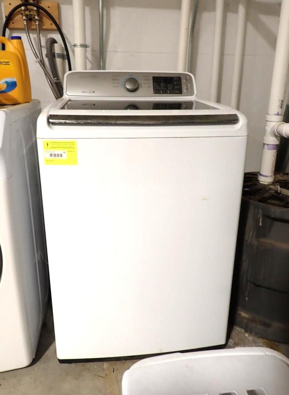 A white top-loading washing machine with a digital control panel sits in a basement near white plumbing pipes.