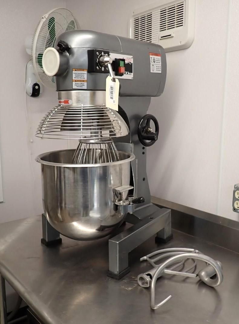 A commercial stand mixer sits on a stainless steel table with extra mixing attachments resting beside the bowl.