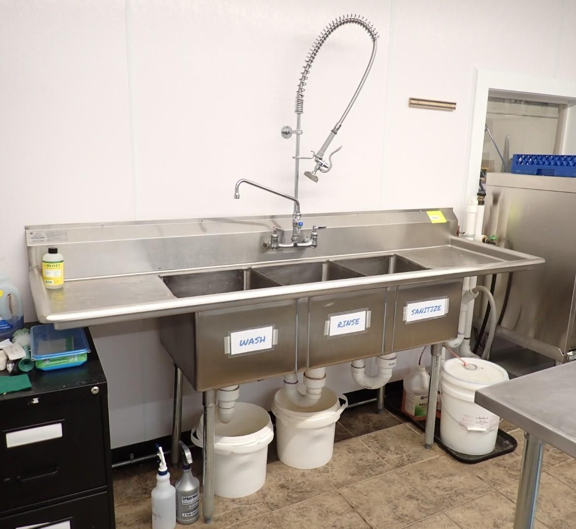 A stainless steel three-compartment commercial kitchen sink with labels, a spray faucet, and buckets underneath.