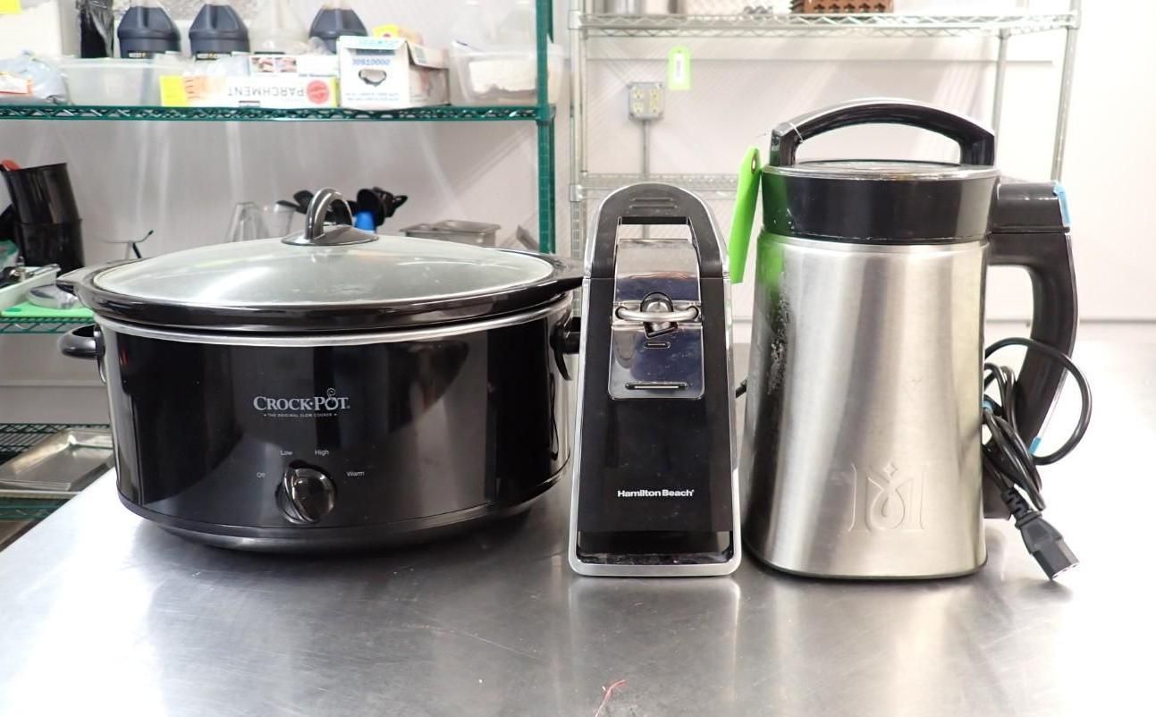 A black slow cooker, a black electric can opener, and a stainless steel electric kettle on a metal surface.