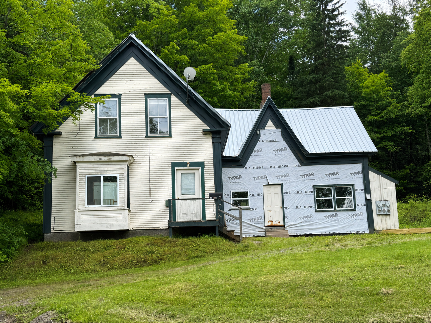 Two small white houses with black trim on a grassy lot, surrounded by trees