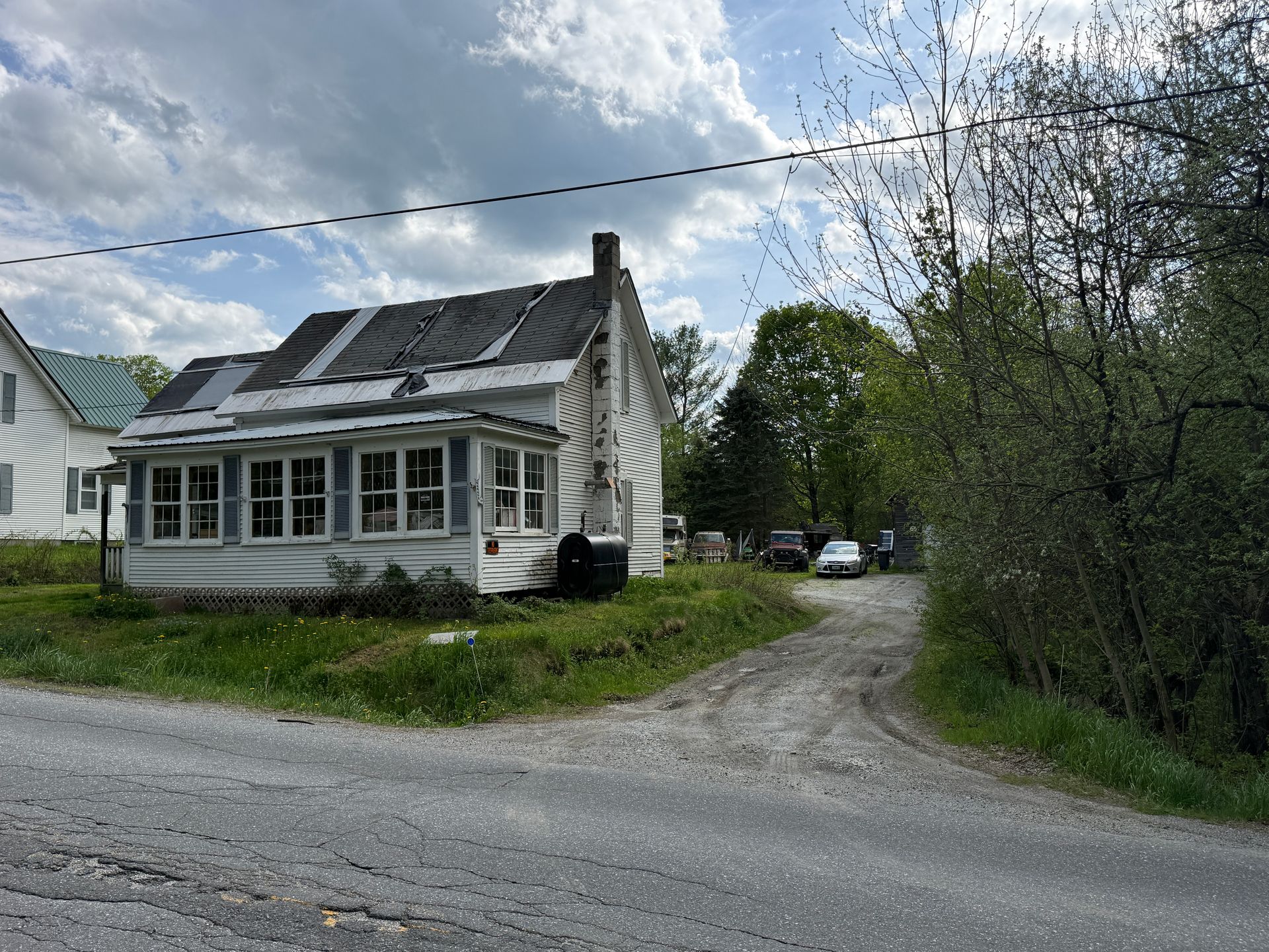 Small white house with a roof beside a gravel driveway and trees under a cloudy sky