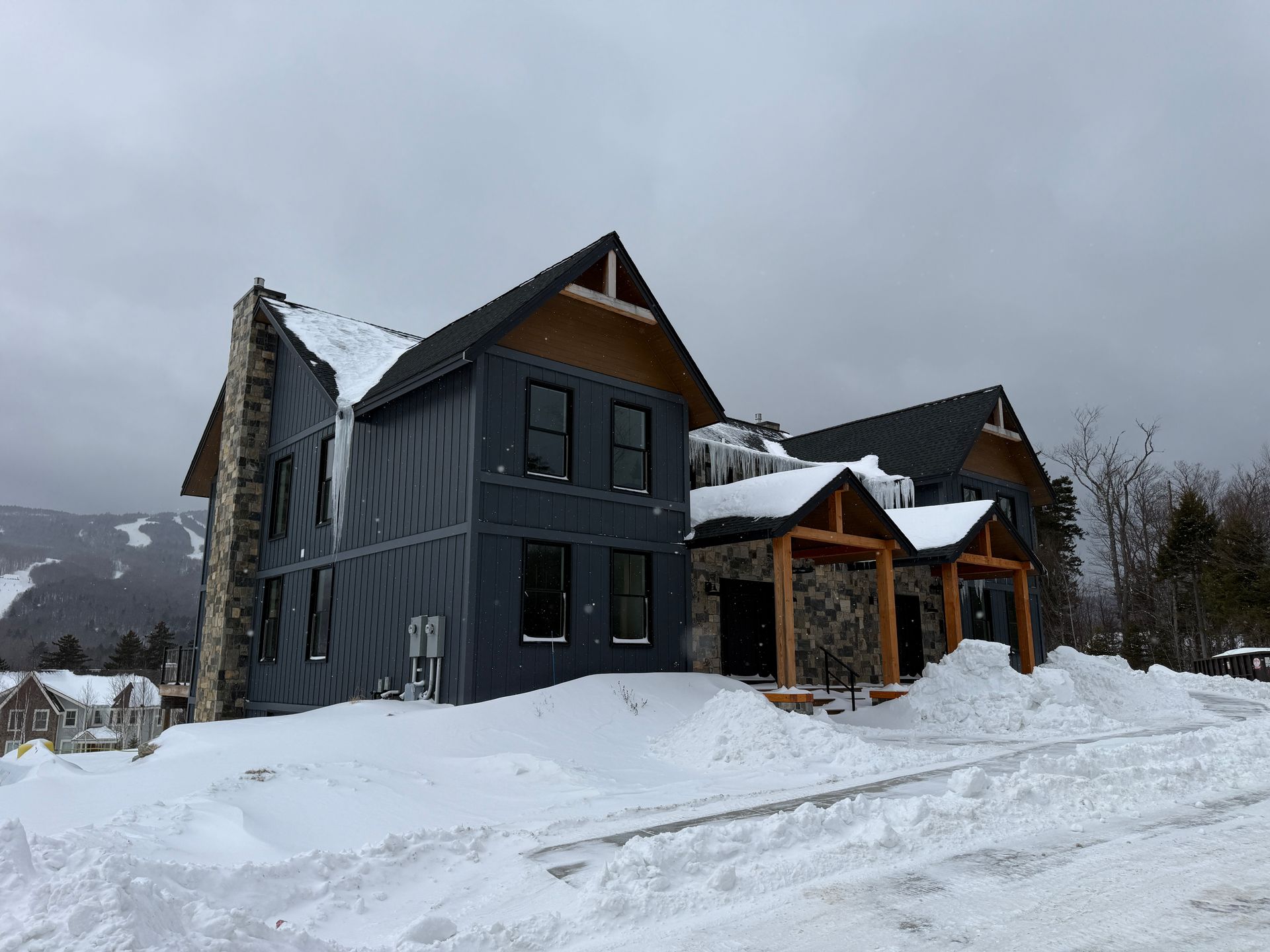 Snow-covered building with dark gray siding, a stone chimney, and wooden accents; Boulder Ridge Foreclosure Property.