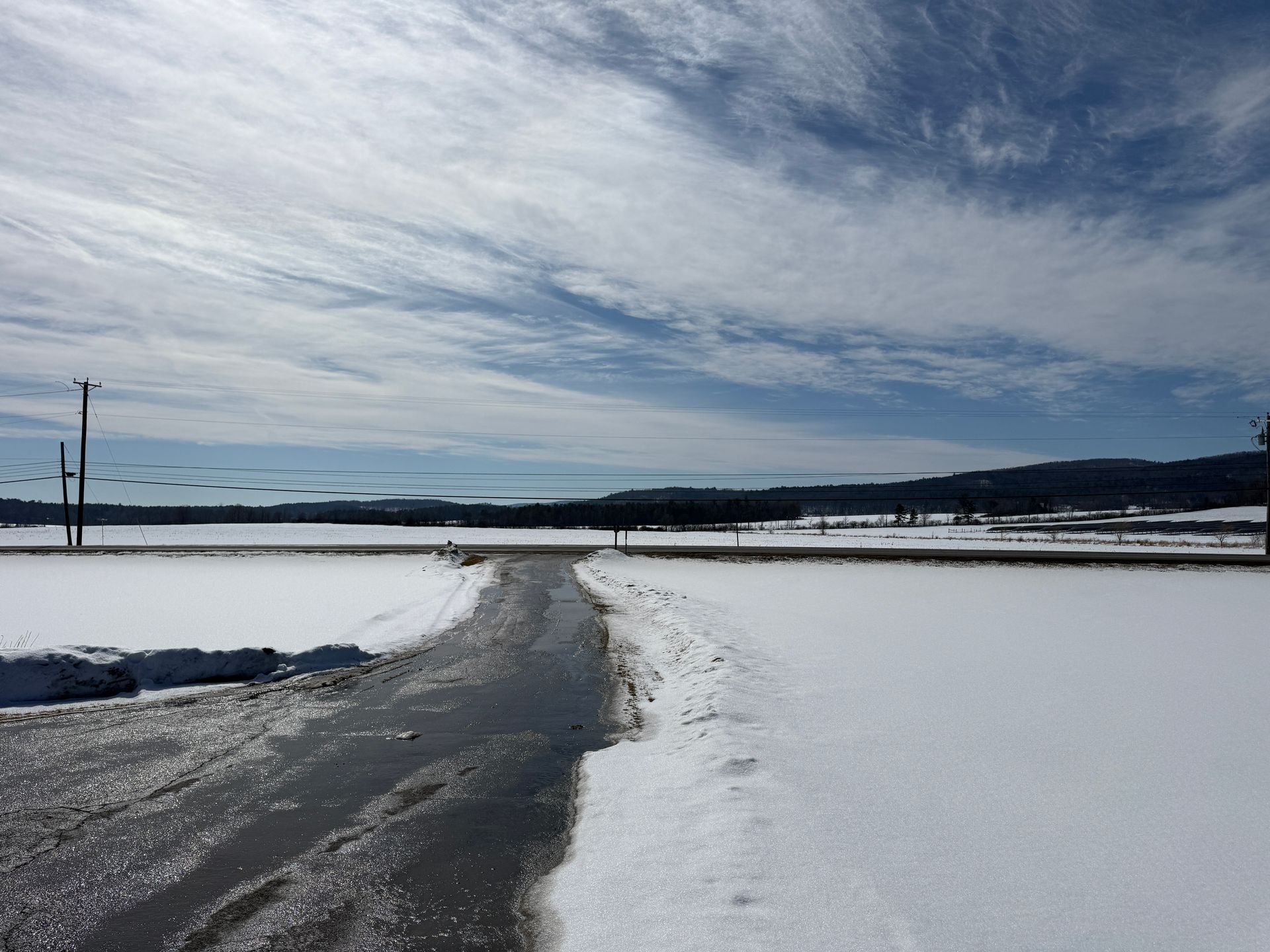Snowy landscape with a partially melted road, blue sky with wispy clouds, and distant trees.