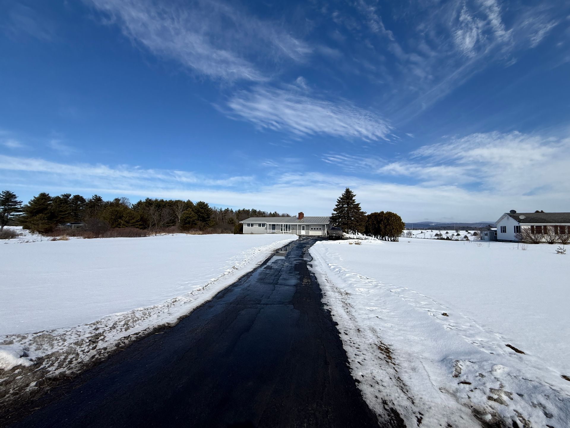 Black asphalt driveway leading to a white house, surrounded by snow under a blue sky.