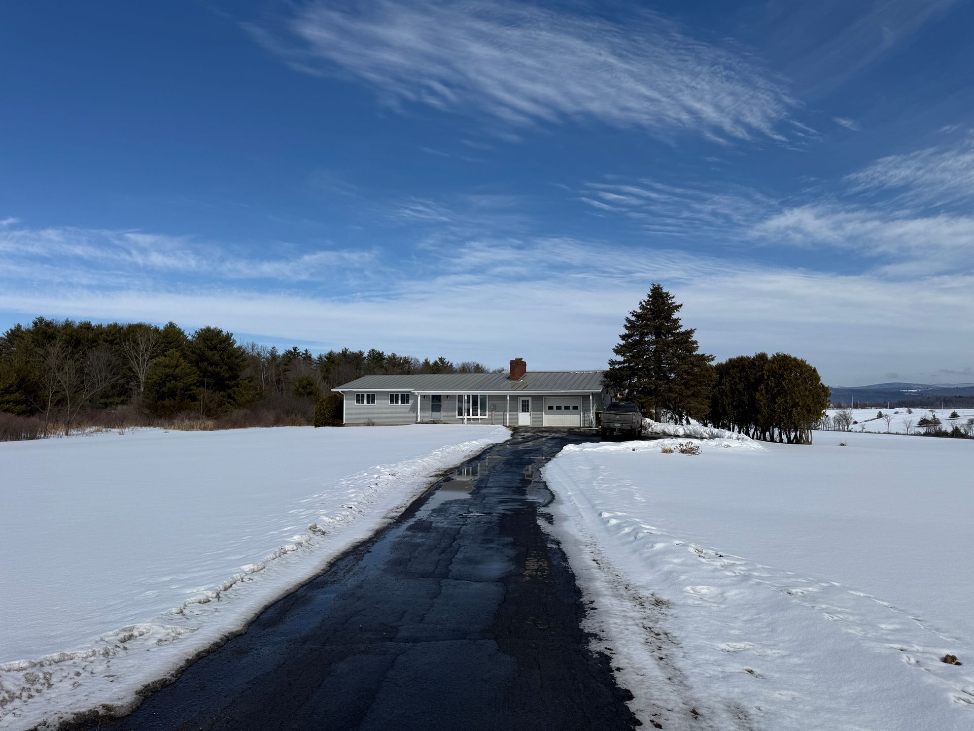 A snow-covered driveway leads to a white ranch-style house under a bright blue sky with wispy clouds.