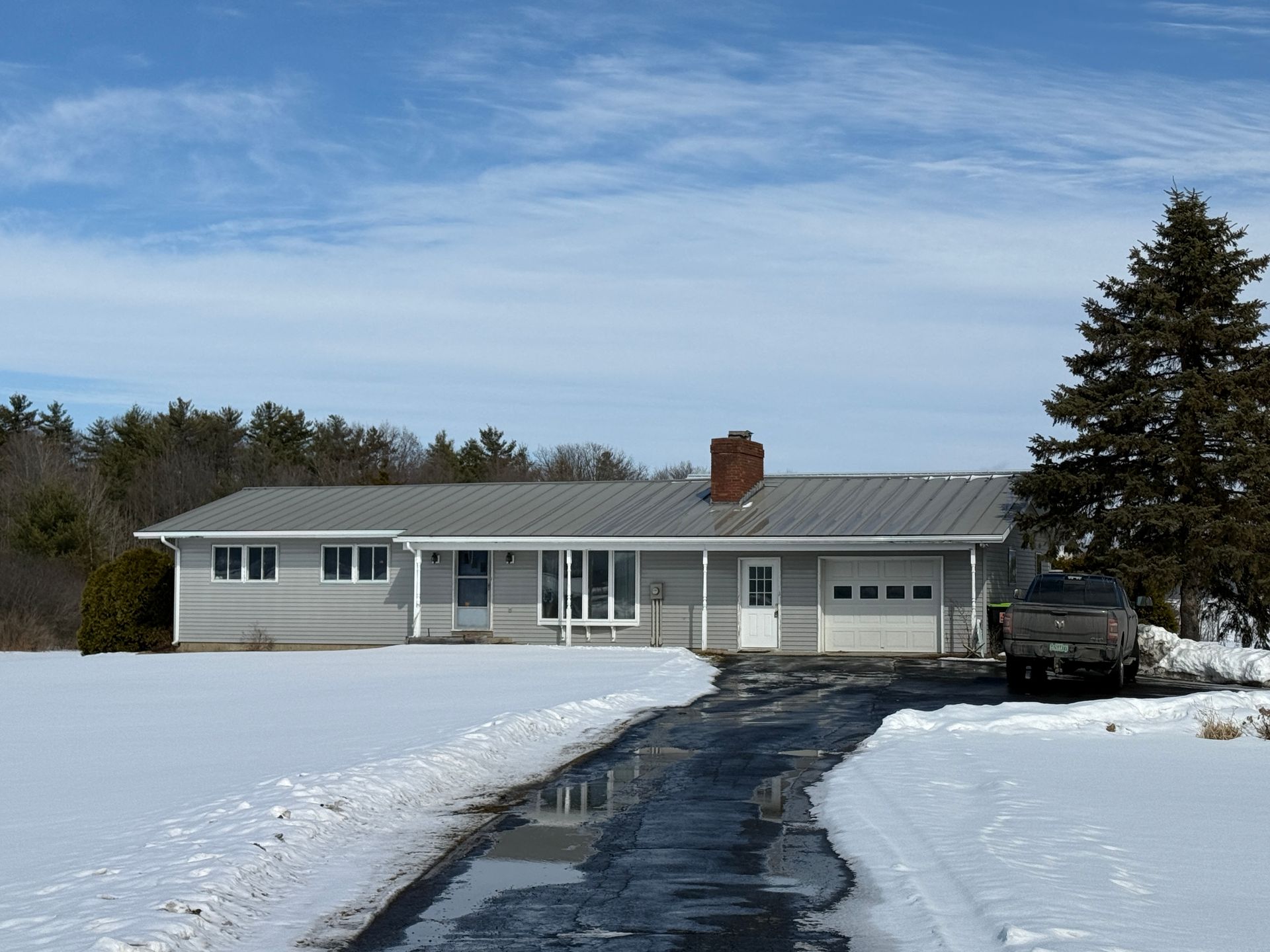 Gray ranch-style house with a snow-covered yard and driveway under a blue sky.
