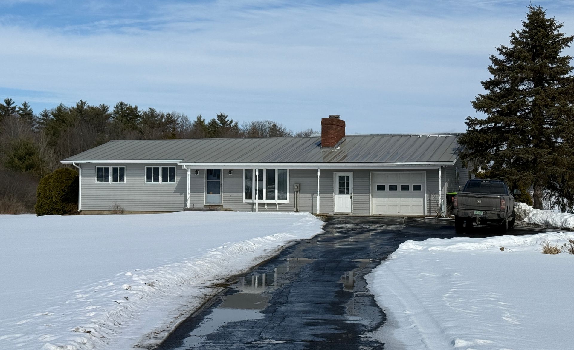 A ranch-style house with a snow-covered yard and partially cleared driveway under a blue sky.