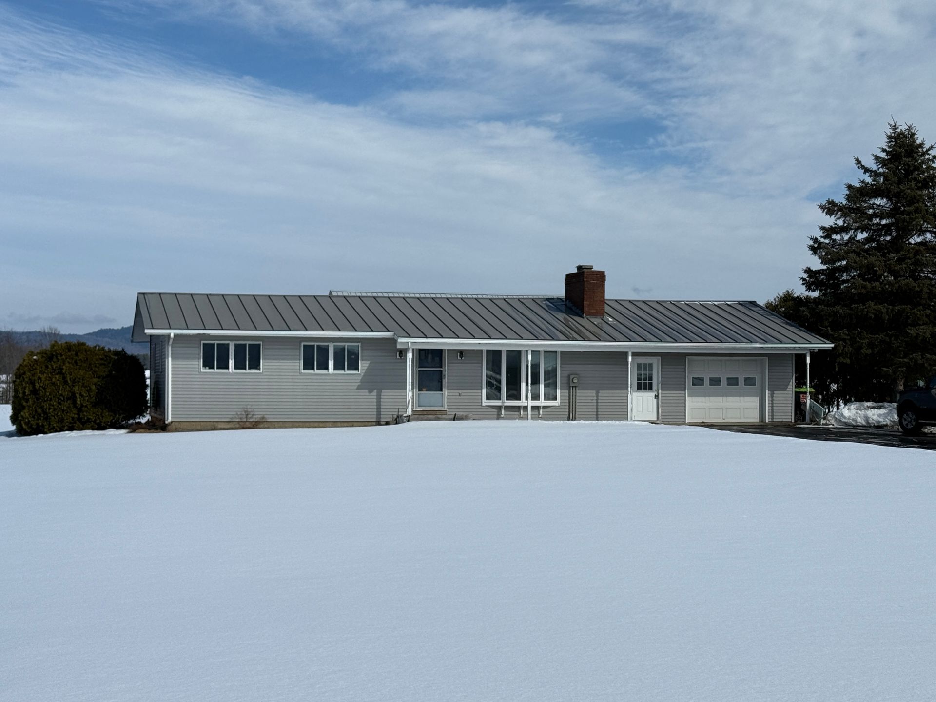 Gray ranch-style house with metal roof, chimney, and attached garage in a snowy field under a cloudy sky.