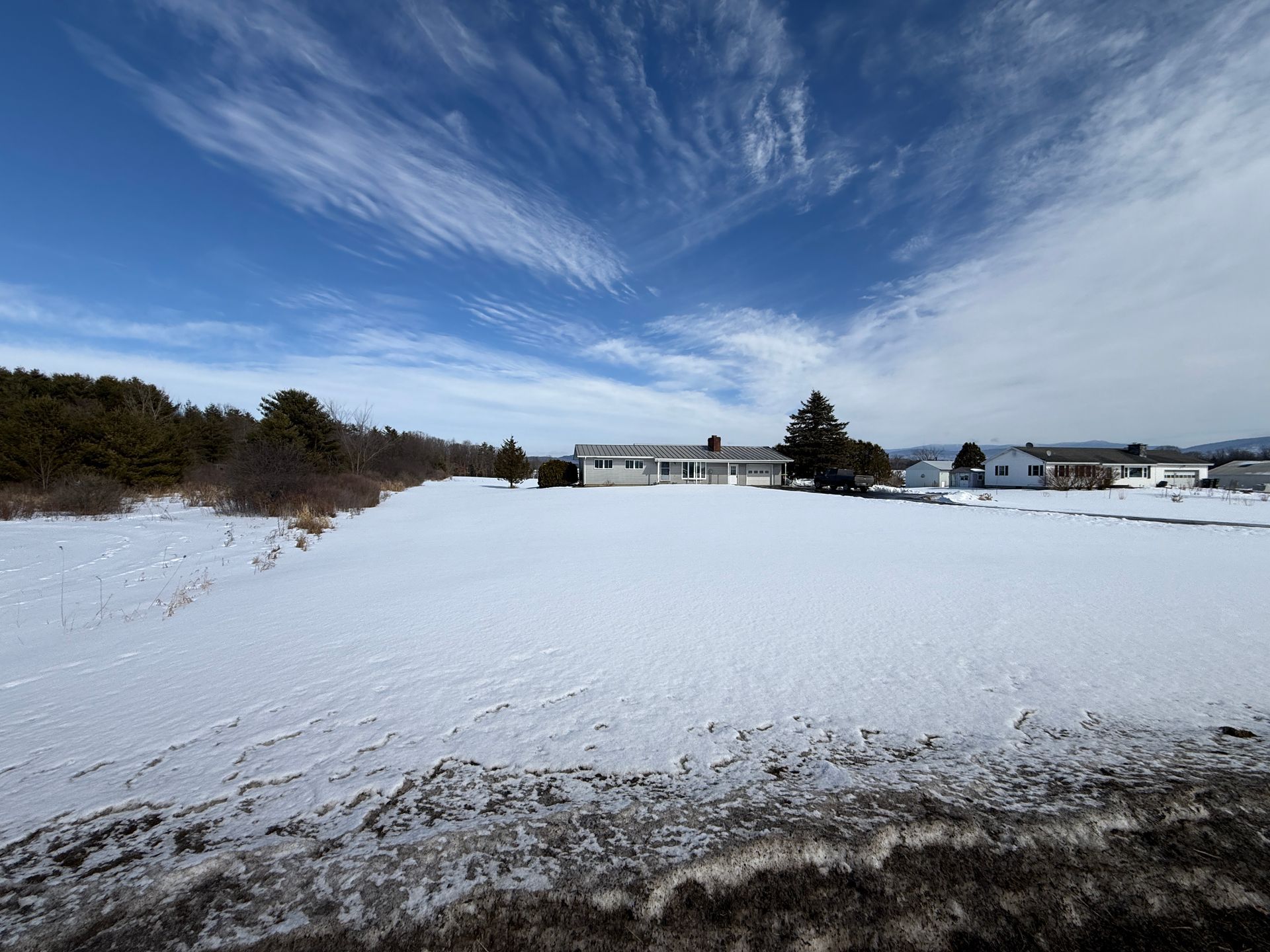 Snow-covered field with houses and trees under a blue sky with streaky clouds.