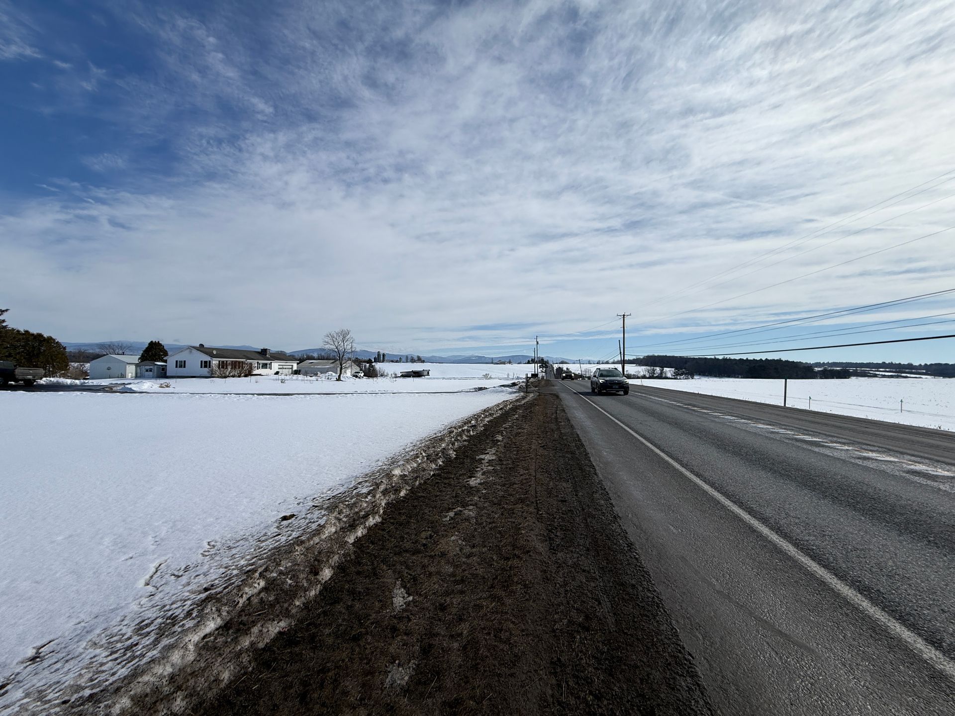 Snowy rural road with car, fields, houses, and cloudy sky.